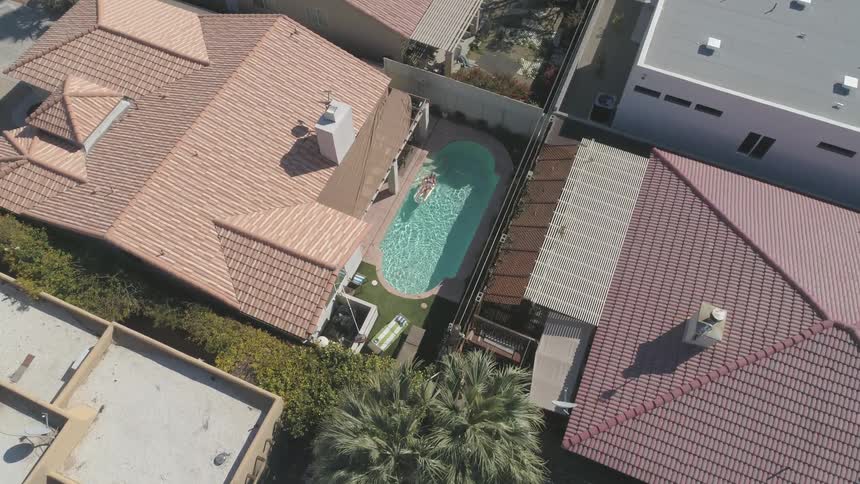 A birds eye view shows a woman floating in an oval pool behind a house