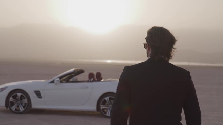 A man in a suit is walking towards a convertible white mercedes benz coupe in a desert