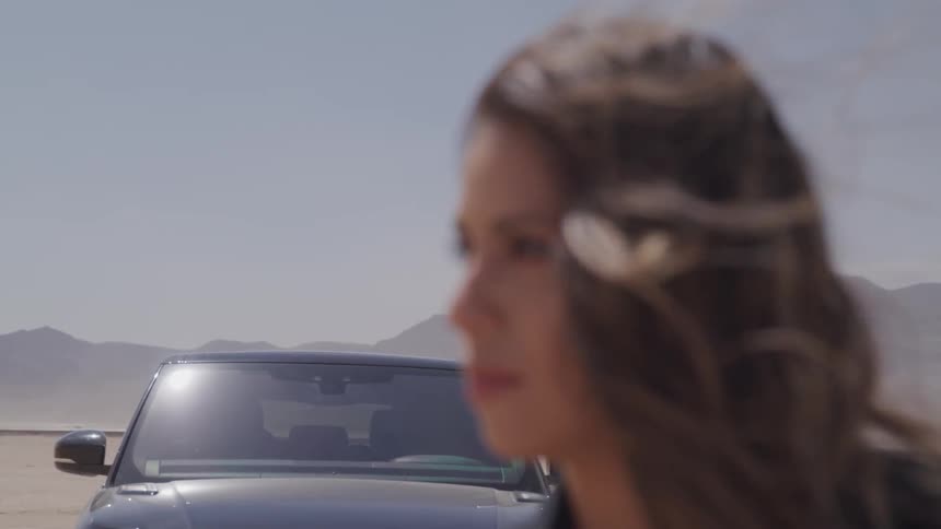A womans hair blows in the wind as she stands in front of a parked car in the desert