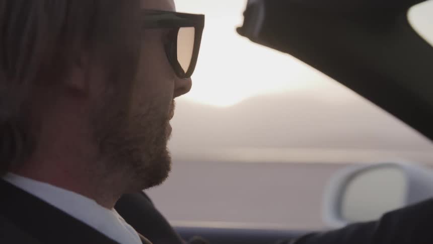 Blonde man wearing sunglasses and suit drives car while sun goes down