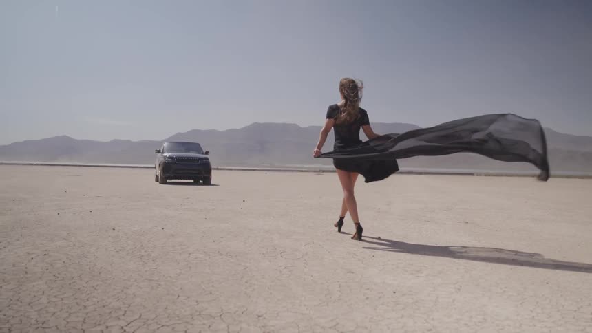 A woman walks towards a car with a cape flowing behind her