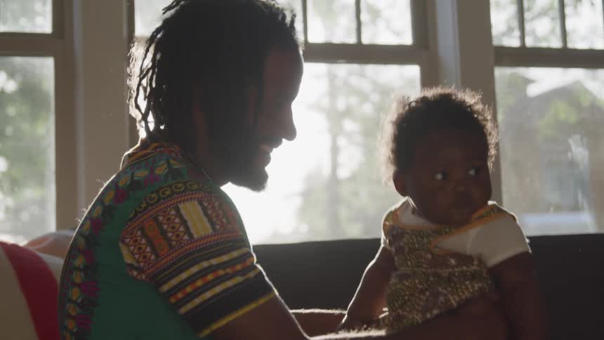 A black man bounces his baby girl on his knee next to a window