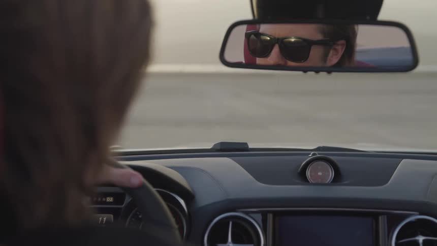 Blonde man wearing sunglasses drives car on dirt road at sunset