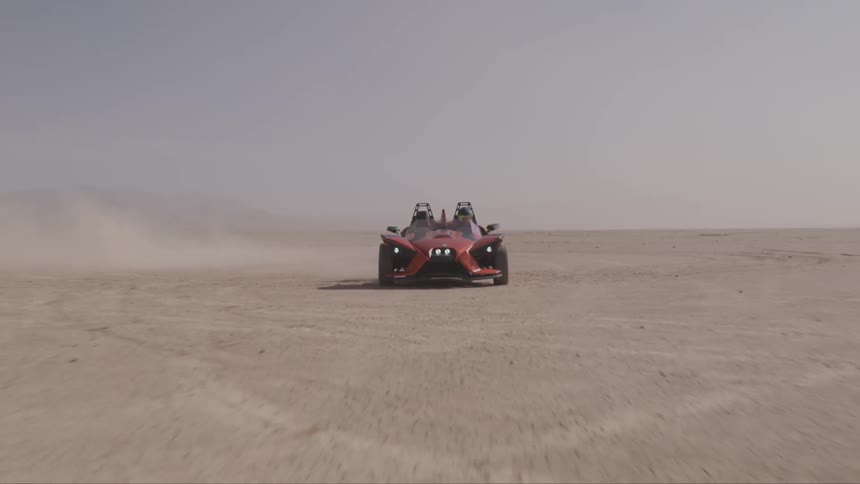 A three wheeled vehicle kicks up dust as it races over cracked dry terrain