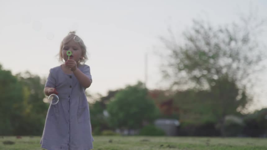 Little blonde girl wearing a dress blowing bubbles at the park