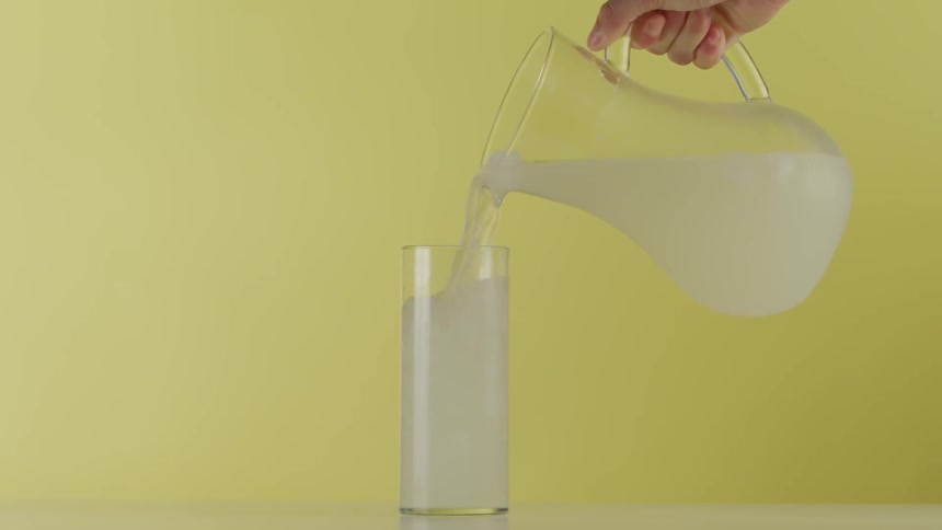 Lemonade is poured from a pitcher into a glass in front of a yellow wall