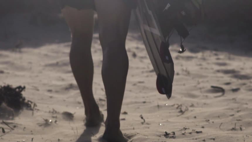 Tight shot of a man's feet and legs as he walks along while carrying a surfboard