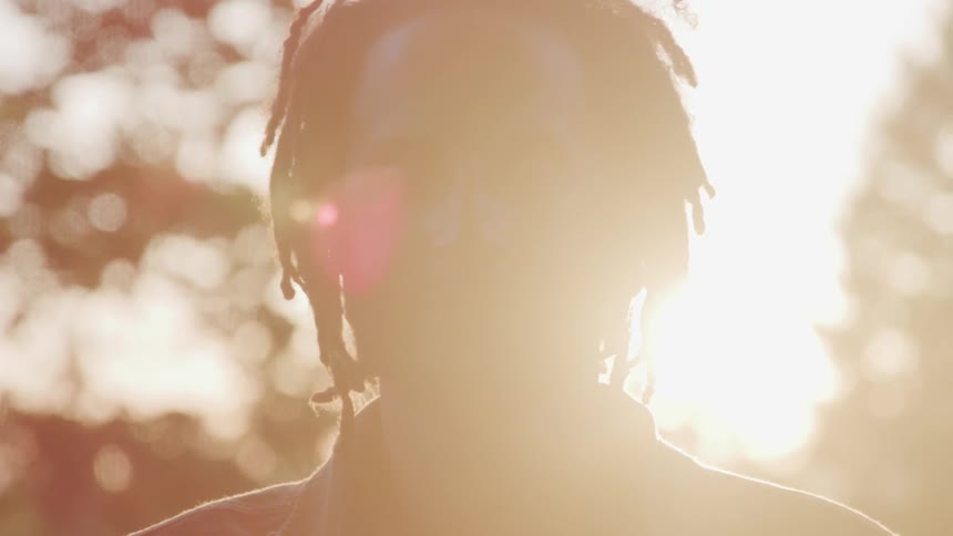 A man with dread locks faces the camera with the sun shining through the trees behind him