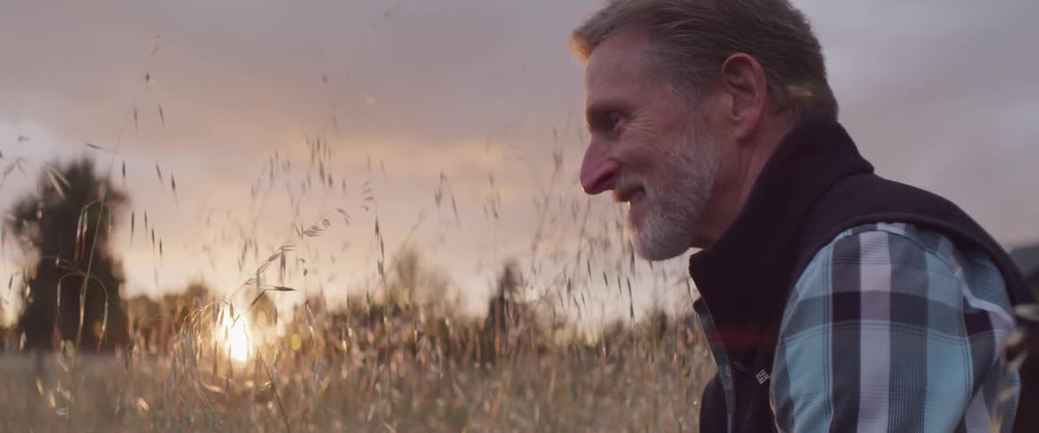 A smiling man sits in a wheat field during sunset