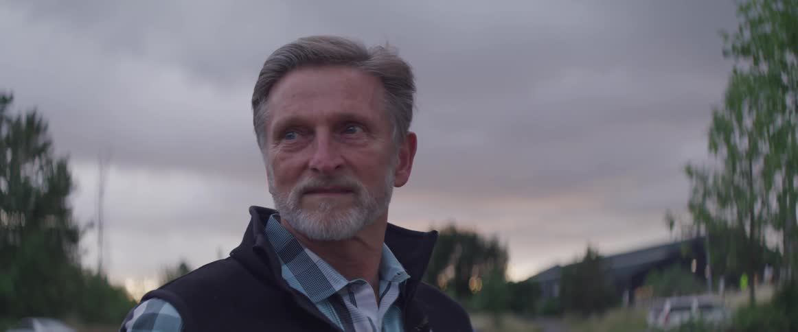 While standing outdoors under a cloudy sky a gray haired and bearded man is looking off in the distance but turns and smiles as he sees something else
