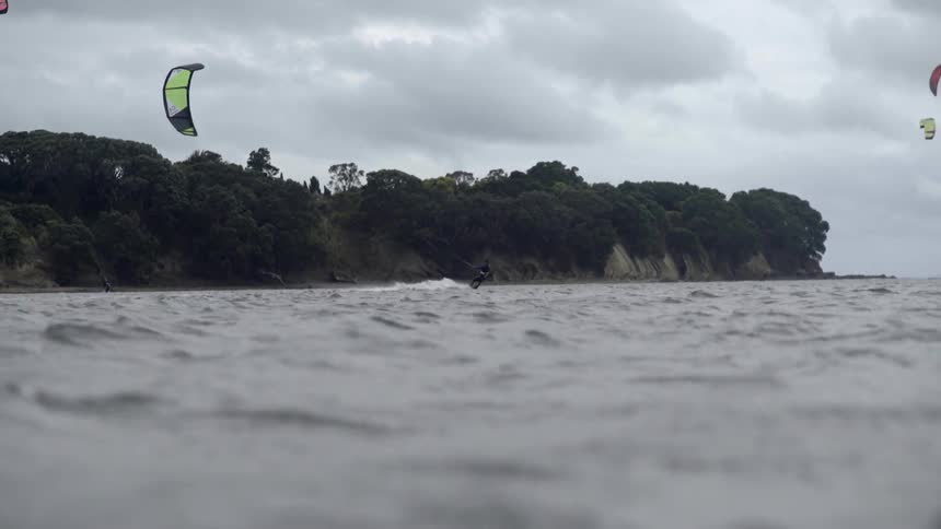 A group of kitesurfers glide over ocean waves beneath a cloudy, overcast sky.