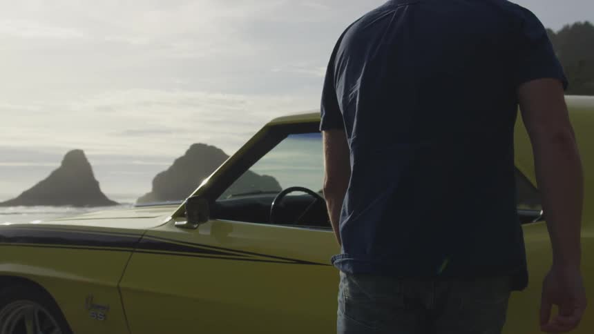 A man walks slowly toward a yellow sports car that is parked near the ocean