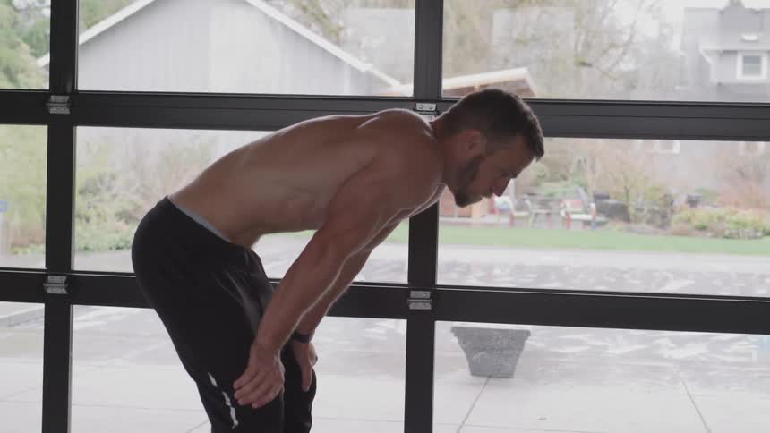 A shirtless man is doubled over and breathing hard while he stands next to a window