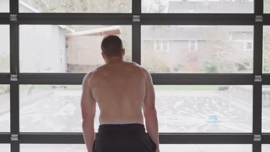 A shirtless man standing in front of a wall of windows bends over with hands on knees to catch his breath