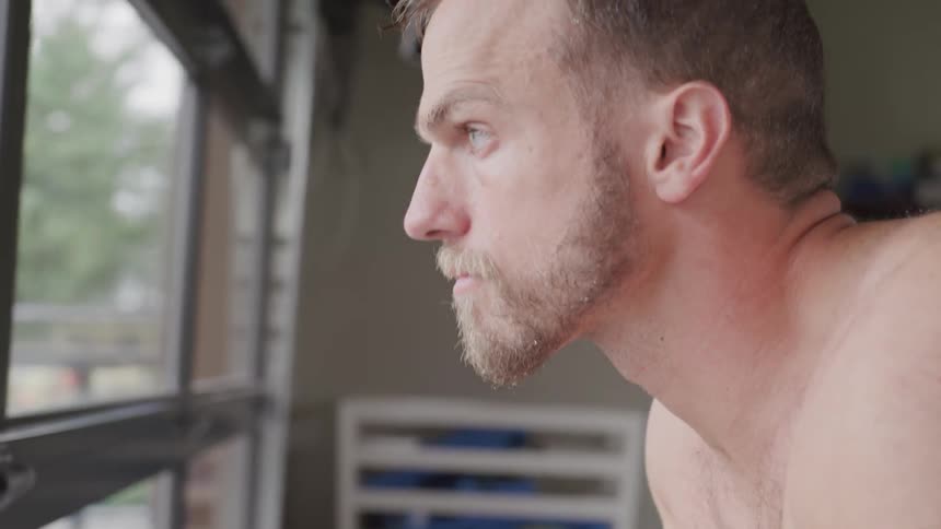 A shirtless man leans forward and stares out a window