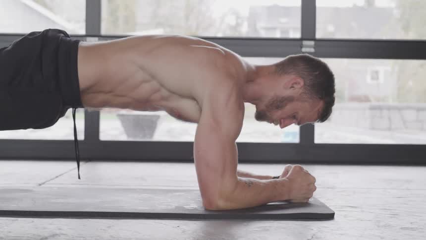 A white male is on a mat doing a forearm plank in front of a wall of windows