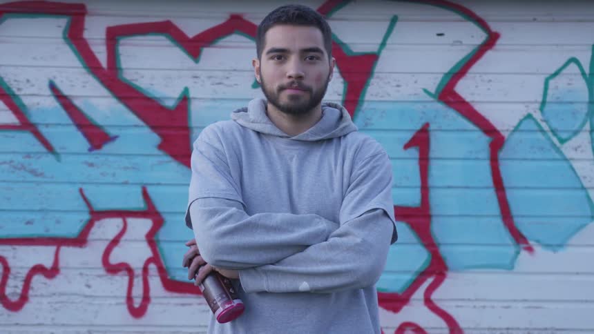 A man holding a can of spray paint stands in front of a graffiti covered wall