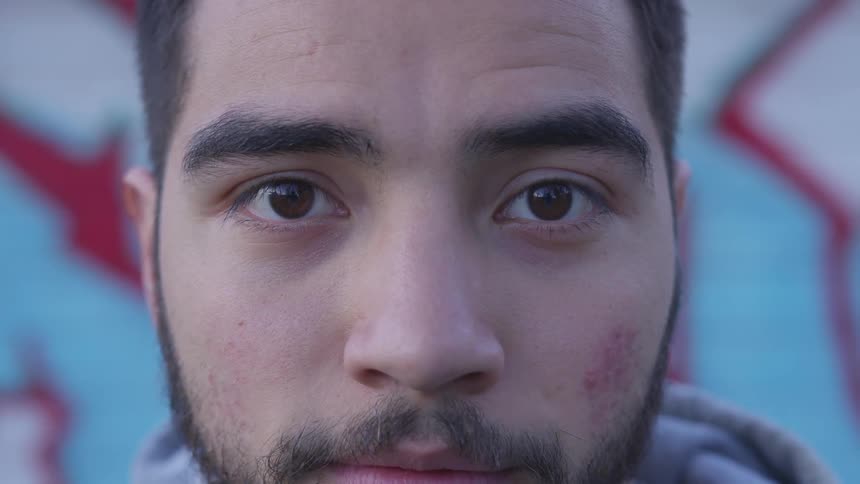 Bearded man stares at the camera in front of a graffiti wall during daylight