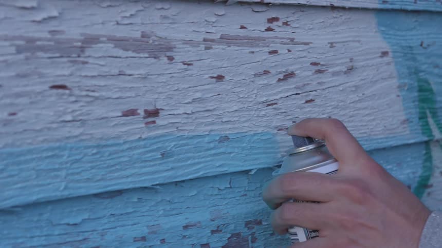 A person spray paints wood siding with blue paint