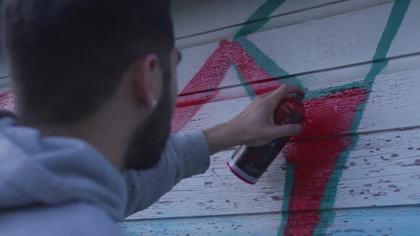 A man uses spray paint to mark red graffiti on the side of a wood structure