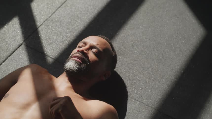 Shirtless black man lies down on the floor and takes deep breaths during daylight