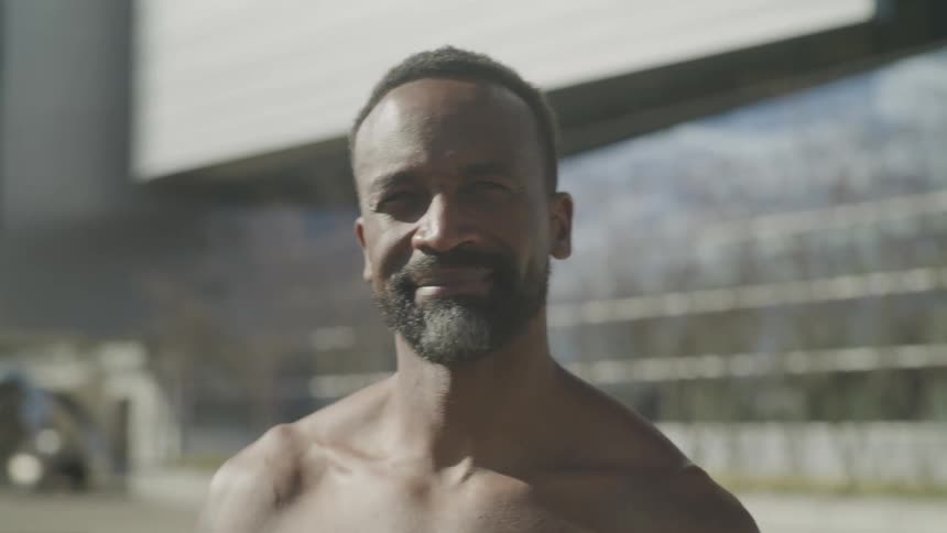 A shirtless black man smiling at the camera while standing outdoors