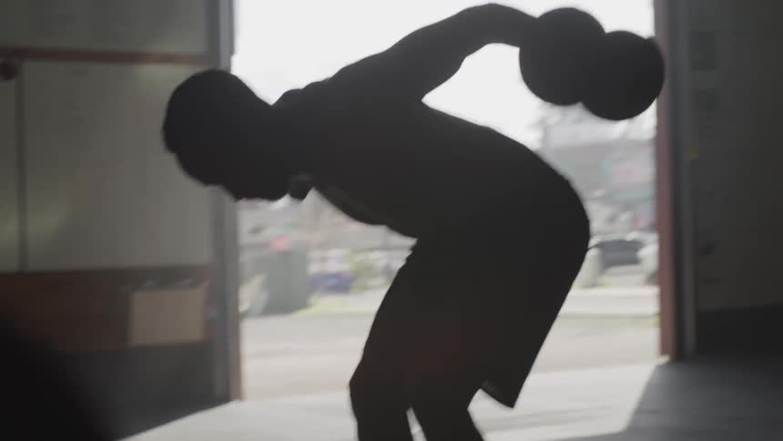 A man is bent over doing dumbbell flies in a building with an open door