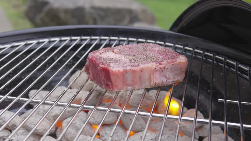 A very thick slab of seasoned steak sits atop a metal rack over a charcoal grill as a small flame flickers below it