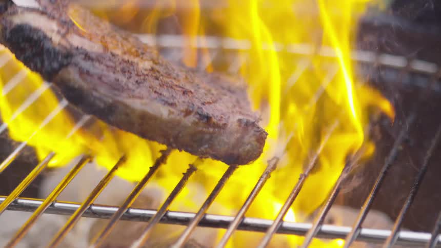 Tongs are used to pick a charred steak up off a flaming grill