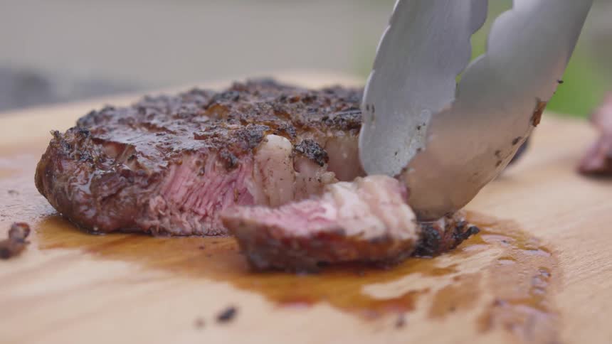 A pair of metal tongs picks up a slice of medium cooked steak from a wood cutting board