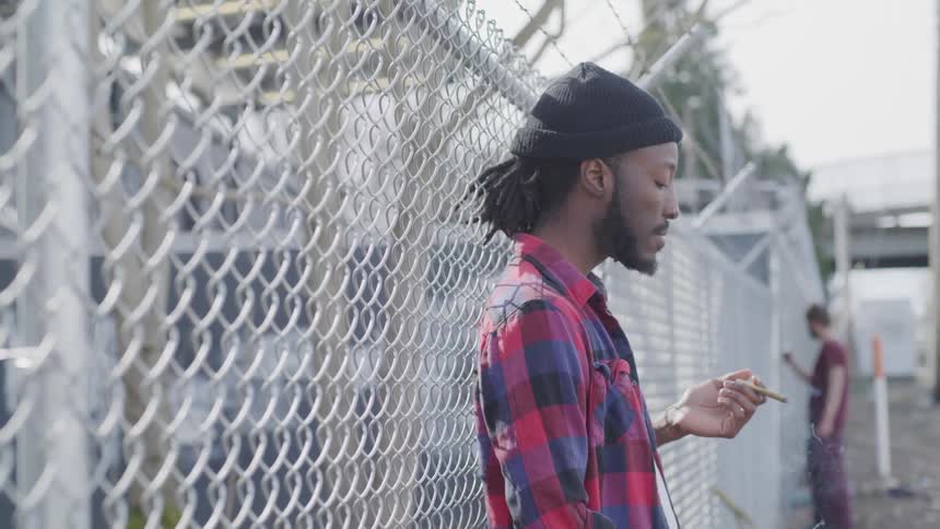A man in a plaid shirt smokes a cigarette while leaning against a chain link fence
