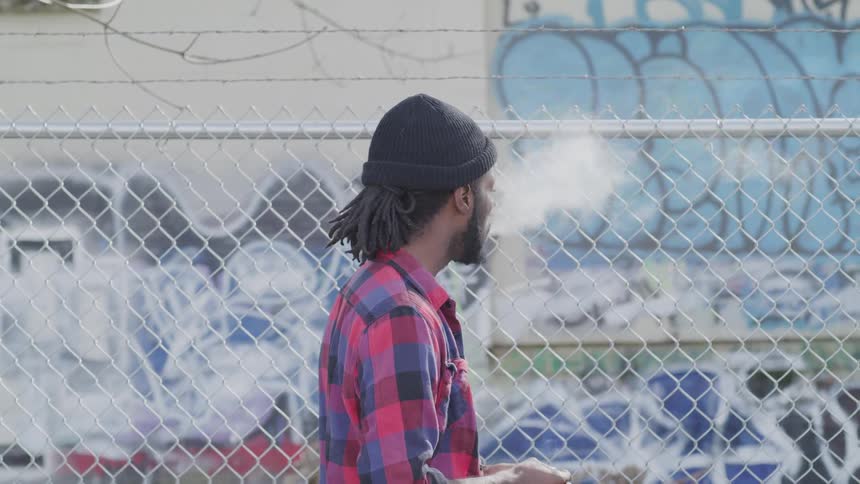 A man smokes while staring at graffiti through a fence