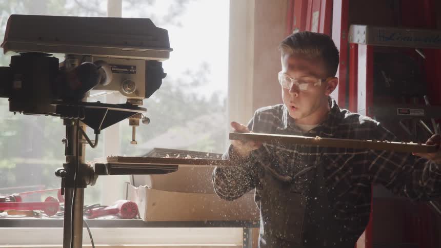 A man blows the sawdust off a board after removing it from a bench drill press