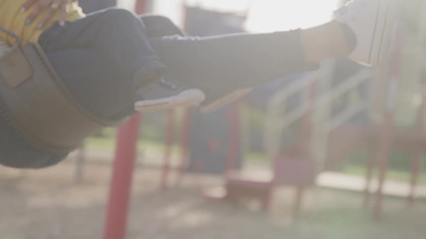 A mom holds her toddler son on her lap as they swing on a swing set in a playground