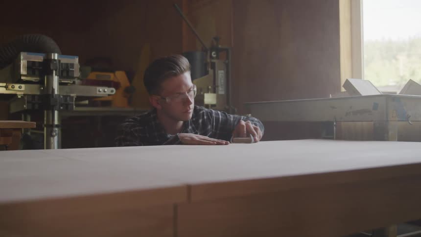 A man wearing goggles in a workshop is sitting eye level with a large wooden table while carefully rubbing sand paper across the smooth top