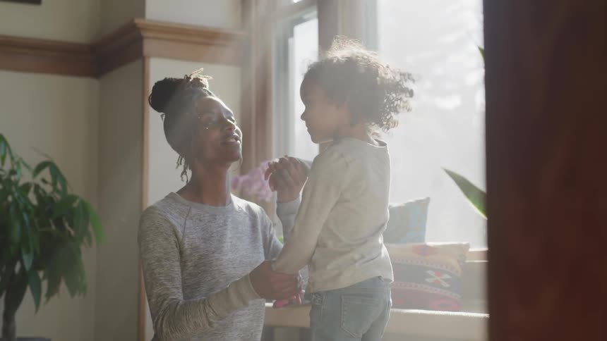 Black woman plays with black toddler girl in a room while the sun streams through the window