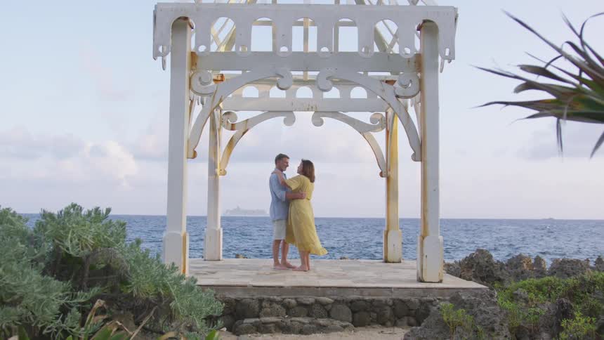 A man and woman hold each other and dance under a pergola near the ocean