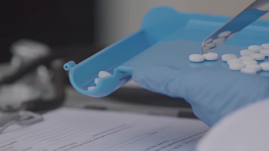 A metal tool pushes white pills around on a blue plastic tray that is held by a gloved hand