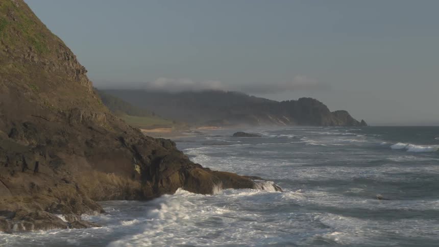 Swift waves are forcefully crashing against the rocky shoreline of a forest covered mountainous coast