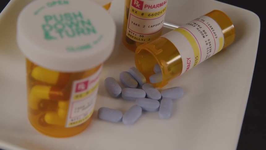 Four bottles of prescription medication are on a white plate and one bottle has fallen over and allowed blue pills to spill out