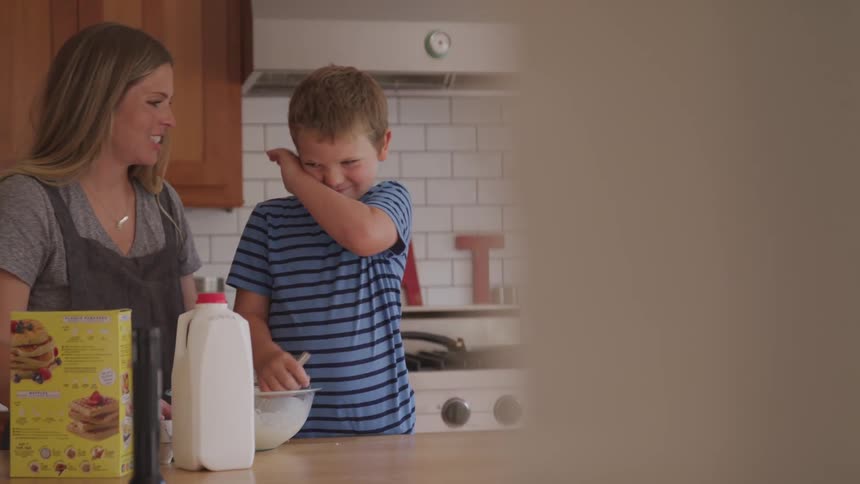 A woman kissing a child while he stirs pancake mix in the kitchen