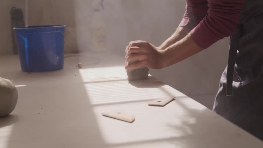 Man wearing black apron kneads dough on the table while the sun streams through window during daylight