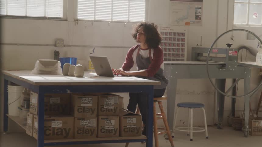 Man sits typing at table in pottery workroom