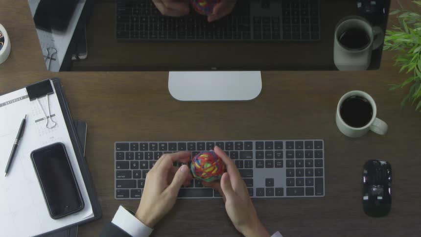 A person sits at a desk and types before picking up a ball of rubber bands playing with it then dropping it onto the desk