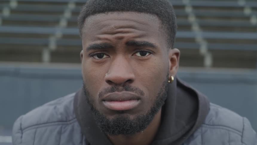 A young black man with a beard turns from facing his right to facing the camera as he sits in front of some blue bleachers