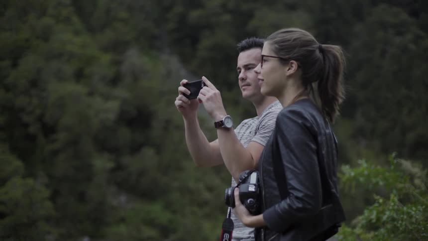 A man and woman are standing by a forest the man is taking photographs