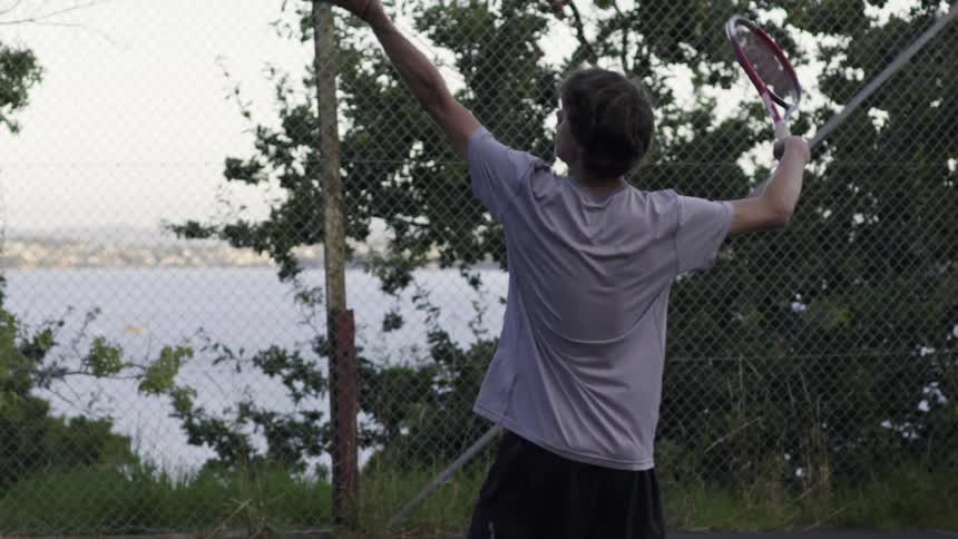 A young man serves a tennis ball on a fenced in court with a harbor or lake in the background