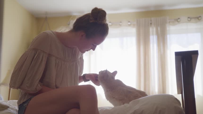A female with her hair up in a bun sits on the edge of a bed petting a cat and scratching it under the chin