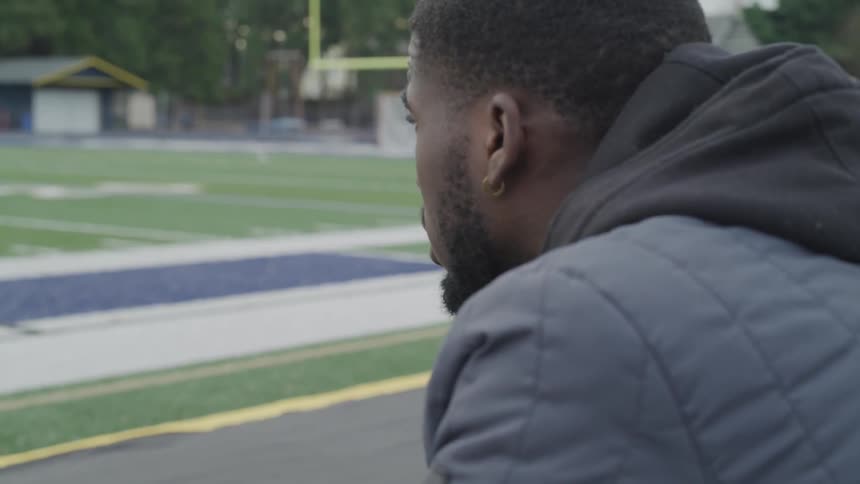 A man with a jacket and hoodie sitting down looking at a football field