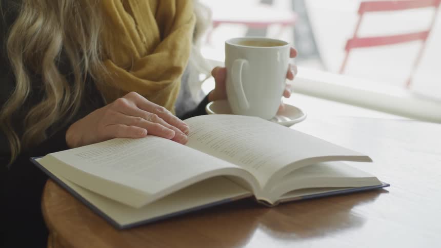 Female sitting at a round table next to the window is sipping coffee while reading a book